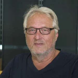 Mann mit grauen Haaren und Brille, der in die Kamera schaut, mit einem neutralen Gesichtsausdruck.