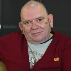 Mann mit Glatze, Gesichtstätowierungen und rotem Cardigan, der in die Kamera schaut.