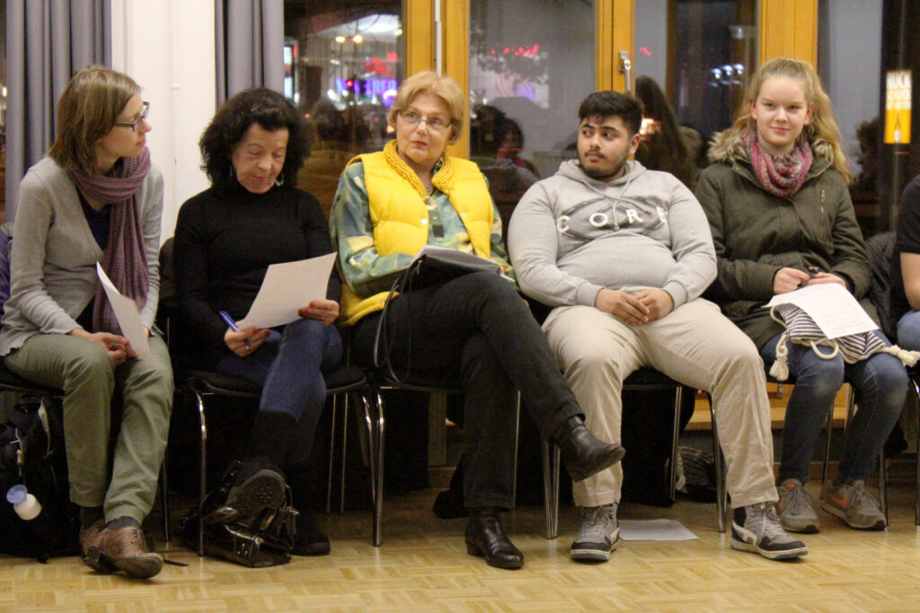 Sechs Personen sitzen in einer Besprechung, einige halten Notizen und schauen aufmerksam.