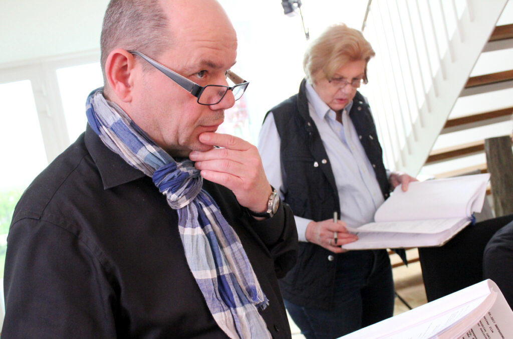 Mann mit Brille und Schal, der nachdenklich wirkt, während eine Frau im Hintergrund Notizen macht.