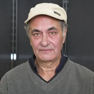 Älterer Mann mit grauem Pullover und beiger Mütze, der direkt in die Kamera schaut.